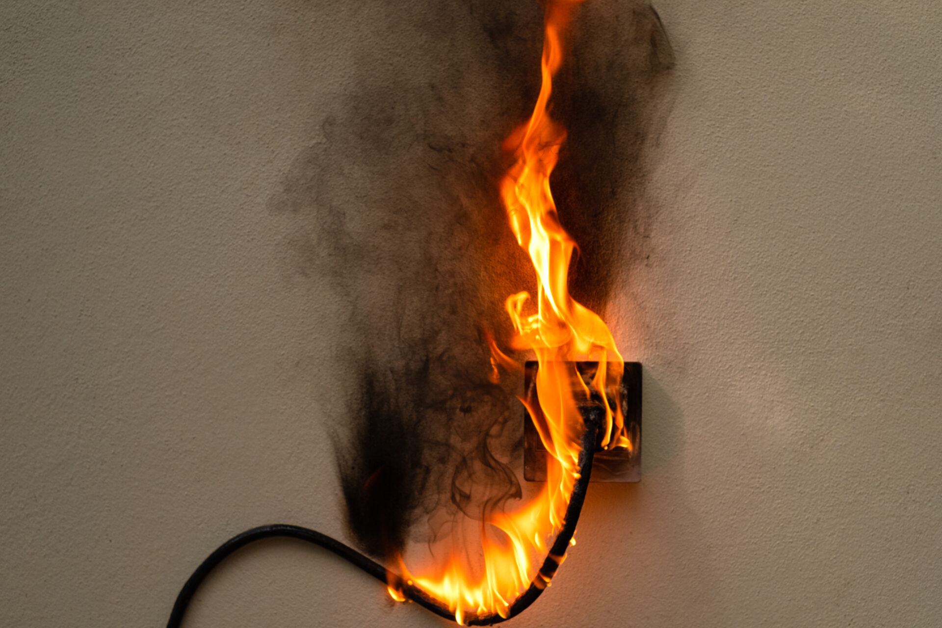 Electric wire plugged into an outlet on a wall. Both are on fire and burning the surrounding wall.